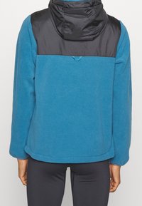Giacca in pile blu con una parte superiore in nylon nero e cappuccio. Presenta una consistenza morbida, maniche lunghe e un orlo rifinito.