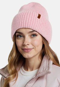 Jasnoróżowy dzianinowy beanie o prążkowanej fakturze. Posiada małą brązową skórzaną metkę. Noszony z jasnoróżową kurtką puchową i białą bluzką.