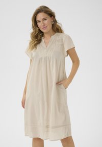 Robe beige à manches courtes avec un décolleté en V, des détails plissés, une coupe ample et des poches, présentant un accent en dentelle au bas. Tissu doux et léger.