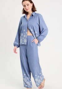 Blouse en lin bleu avec un devant à boutons et une broderie florale à l'ourlet, associée à un pantalon ample assorti. Tissu léger, coupe décontractée.