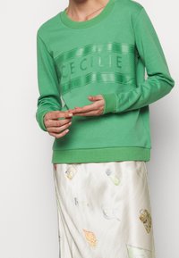 Felpa verde con colletto e polsini a costine, che presenta una fascia con logo stampato lucido che recita "CECILIE." Abbinata a una gonna in seta con fantasia.