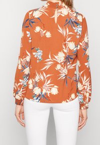 Blusa floreale in arancione bruciato con fiori bianchi e blu. Presenta un colletto alto, chiusura con bottoni e maniche arricciate. Tessuto dalla consistenza liscia.