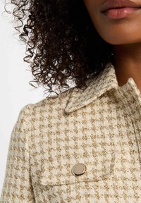 Veste en tweed crème et beige avec un col, motif texturé et un accent en métal argenté sur le côté gauche.