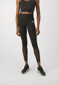 Leggings sportivi neri con pannelli in rete, vita alta e dettagli testurizzati, abbinati a un reggiseno sportivo nero e scarpe.