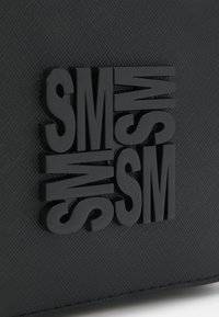 Schwarze Tasche mit erhabenen, strukturierten Buchstaben "SM" in einer fetten, modernen Schriftart. Das Material wirkt langlebig mit einem dezenten Muster und glatter Oberfläche.