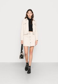 Veste beige raccourcie avec deux poches avant, col roulé noir, jupe beige à boutons sur le devant, et bottines noires avec un matériel circulaire.