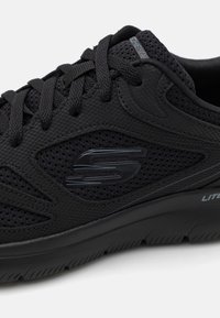 Närbild av en svart sportskshoe med nätmaterial, texturerad sula och en blank logotyp på sidan som framhäver den sportiga designen.