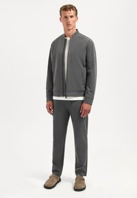 Grijze bomberjack met rits en een ronde hals, gecombineerd met bijpassende broek. Gestreepte wit-grijze t-shirt eronder. Beige schoenen.