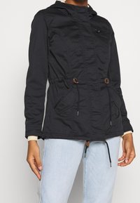 Chaqueta negra con capucha, con cordones elásticos, bolsillo con cremallera y bolsillos laterales. Presenta una tela texturizada y puños acanalados para mayor comodidad.