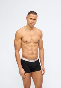 Άνδρας χωρίς πουκάμισο με κοντά μαλλιά, φορώντας μαύρα μποξεράκια Tommy Hilfiger, στέκεται μπροστά σε απλό λευκό φόντο, κοιτάζοντας προς τα εμπρός.