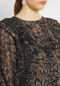 Haut imprimé serpent marron et noir avec des détails de col volanté. Tissu transparent avec une texture lisse. Accessoire : grande boucle d'oreille florale rose.