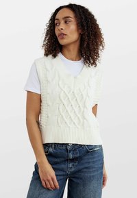 Cremefarbener, ärmelloser Pullover aus Kabelstrick mit V-Ausschnitt und geripptem Saum, kombiniert mit einem weißen T-Shirt und blauen Jeans.