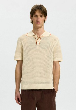 Ung mann med beige kortermet strikket polo med oransje kant og brune kordfløyelsbukser, stående foran en ensfarget hvit bakgrunn.