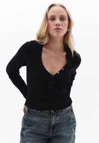 Maglietta nera a maniche lunghe con scollatura a V, dotata di tre bottoni sul petto. Abbinata a jeans in denim blu larghi.