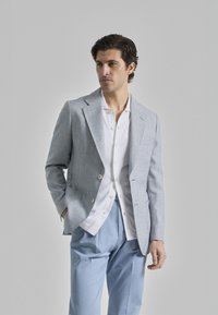 Blazer a cuadros azul claro con solapas de punta, botones dorados y una camisa blanca debajo, combinado con pantalones azul claro.