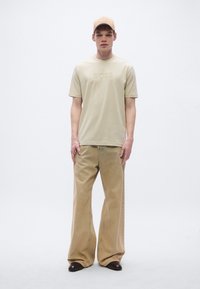 Bézs BOSS póló dombornyomott logóval, bő szárú bézs kordbársony nadrággal párosítva. A modell egy hozzáillő bézs sapkát visel. Simogató tapintású, laza szabású.