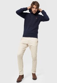 Marineblauwe hoodie met een gestructureerd bovenstuk, een gladde ondersectie, verstelbaar trekkoord en een klein logo-accent. Gecombineerd met beige broeken en bruine laarzen.