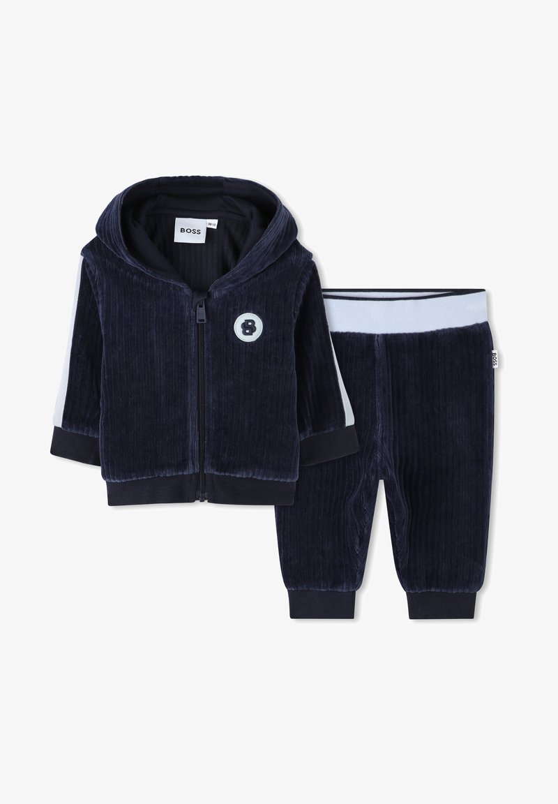 Marino modra kordna zip-up kapuca z rebrastimi manžetami in okroglo logotipom. Ujemajoče se trenirke z elastičnim pasom in enobarvnimi akcenti.