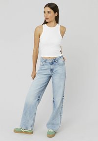 Débardeur blanc côtelé, jeans taille haute bleu clair à jambes larges avec décoloration et distresses, associés à des baskets vertes avec des semelles beiges.