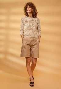 Femme aux cheveux bouclés portant un top en dentelle beige, un short beige longueur genou et des sandales slides tissées noires, debout devant un fond beige.