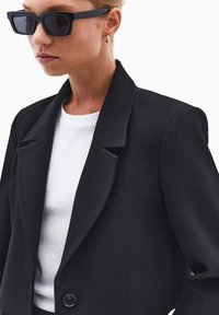 Blazer nero con colletto strutturato e chiusura a un bottone, abbinato a una camicia bianca a girocollo e occhiali da sole neri rettangolari.