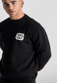 Mann in einem schwarzen Pullover mit einem weißen Logo auf der Brust, stehend vor einem schlichten hellgrauen Hintergrund.
