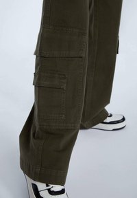 Cargo broek in olijfgroene katoen met meerdere zijzakken, brede pijpen en een rechte snit, gecombineerd met zwart-witte sneakers.