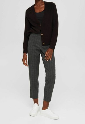 Person wearing black buttoned cardigan, black top, gray checkered pants, and white sneakers standing against plain background.