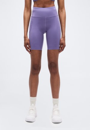 Vrouw die paarse sportshorts met hoge taille en zijzak draagt, witte sneakersokken en witte sneakers, staand tegen een effen achtergrond.