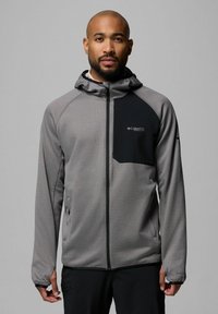 Szara, strukturalna kurtka z czarną kieszenią na piersi i kapturem. Posiada zamek błyskawiczny z przodu oraz elastyczne mankiety, wykonana z oddychającego materiału.