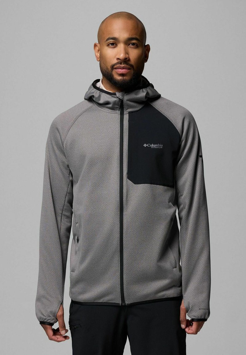 Szara, strukturalna kurtka z czarną kieszenią na piersi i kapturem. Posiada zamek błyskawiczny z przodu oraz elastyczne mankiety, wykonana z oddychającego materiału.