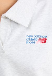 Šedé polo tričko s plochým golierom a vyšívaným nápisom "new balance athletic shoes" v modrej a červenej farbe na ľavej strane. Mäkká textura.