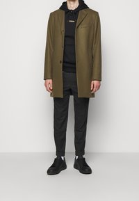 Abrigo de lana color oliva con cierre de un solo botón, combinado con una sudadera con capucha negra, pantalones grises de corte entallado y zapatillas negras. Ajuste sencillo y estructurado.