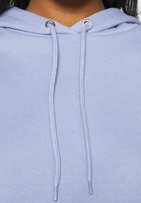 Vaaleansininen huppari, jossa on teksturoitu kangas, varustettuna nyörillä, jossa on hopeiset metallireiät, ja yksinkertainen muotoilu.