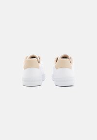 Weiße Sneaker aus glattem Leder mit einem kontrastierenden beigen Fersenpatch und strukturiertem Akzent. Logo auf der Ferse geprägt.