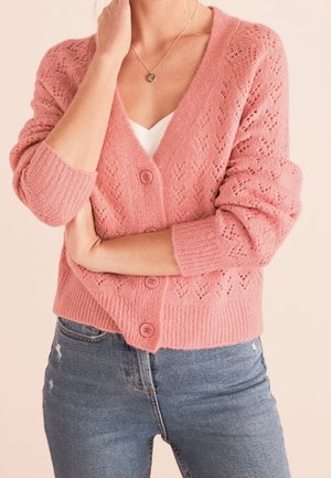 Žena v růžovém pleteném svetru s knoflíky přes bílé tričko, v modrých džínách, se zkříženýma rukama a jednou rukou se dotýká krku.