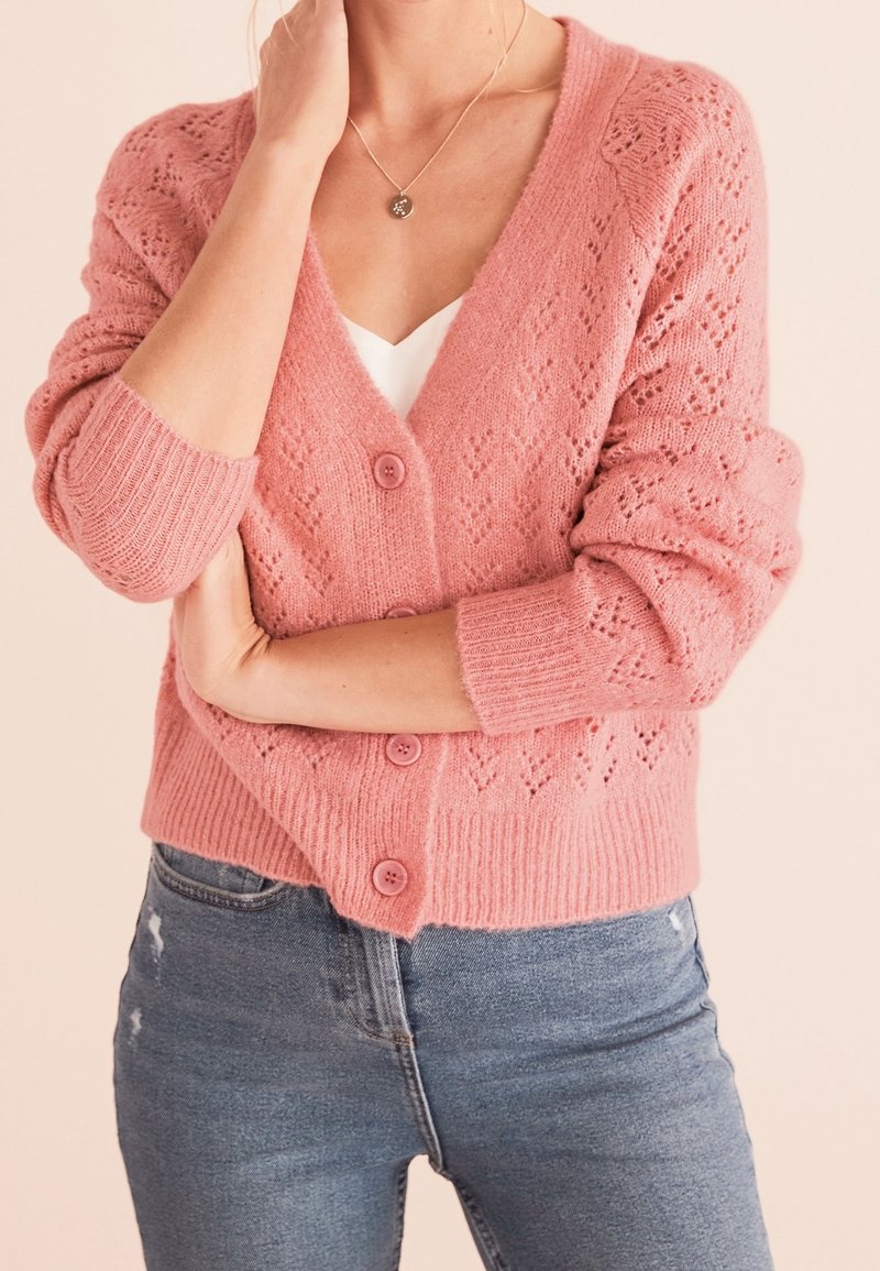 Žena v růžovém pleteném svetru s knoflíky přes bílé tričko, v modrých džínách, se zkříženýma rukama a jednou rukou se dotýká krku.