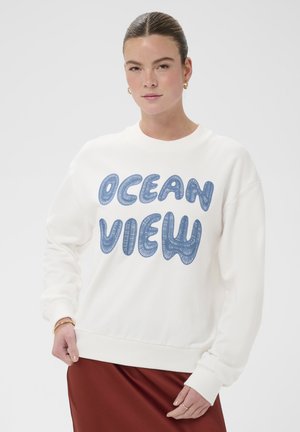 Młoda kobieta w białej bluzie z niebieskim napisem „OCEAN VIEW” i rudą spódnicą, pozująca na jednolitym jasnym tle.