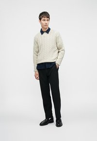 Pull en maille beige porté sur une chemise à col bleu marine, associé à un pantalon noir et des chaussures noires brillantes. Design simple et texturé.