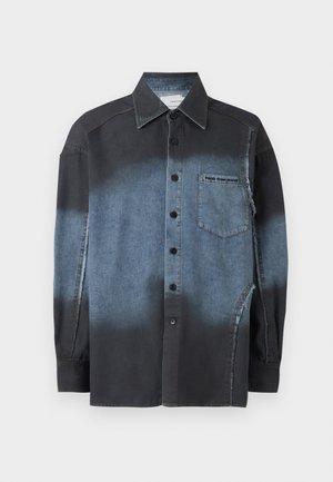 Νεαρός με κατσαρά μαλλιά που φοράει ξεθωριασμένο μαύρο και γκρι πουκάμισο με κουμπιά, μαύρη πλισέ φούστα, μαύρες κάλτσες και μαύρα αθλητικά παπούτσια, όρθιος.