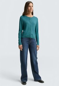 Türkisfarbener langärmliger Cardigan mit rundem Ausschnitt und Knopfverschluss vorne, kombiniert mit blauen Jeans mit weitem Bein und schwarzen Schuhen.