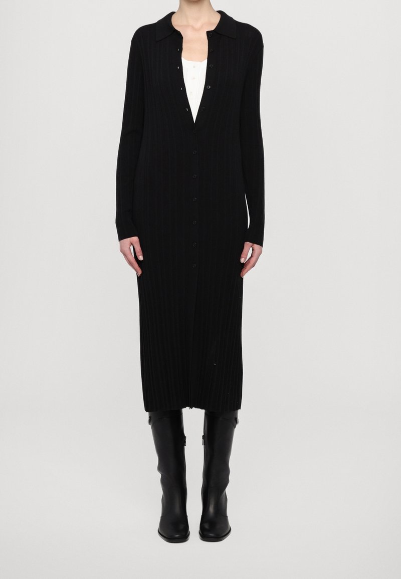 Femme portant une longue robe noire boutonnée sur un top blanc et des bottes noires montantes, debout devant un fond clair uni.
