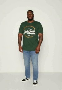 T-shirt verde-escuro com texto gráfico branco, jeans de ganga e ténis pretos com detalhes brancos, em pé contra um fundo simples.