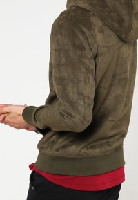 Felpa verde oliva con una texture morbida, dotata di orlo e polsini a costine aderenti. Il design include un cappuccio e dettagli di cucitura discreti.