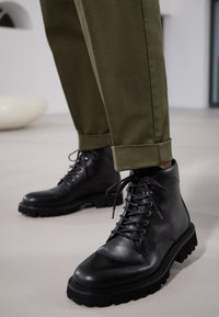 Bottes en cuir noir avec une semelle en caoutchouc épaisse, présentant un design à lacets, une texture lisse et un nez renforcé. Portées avec un pantalon olive retroussé.
