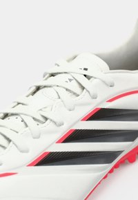 Λευκά ποδοσφαιρικά παπούτσια με λείο δερμάτινο επάνω μέρος, μαύρες και φωσφορίζουσες ροζ λεπτομέρειες, και διακριτό σχέδιο με τρεις ρίγες. Υφασμένη σόλα με καρφιά.