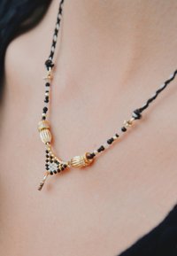 Collier de perles dorées et noires avec un design triangulaire, orné d'accents décoratifs et d'une corde tissée, mettant en valeur des motifs complexes.