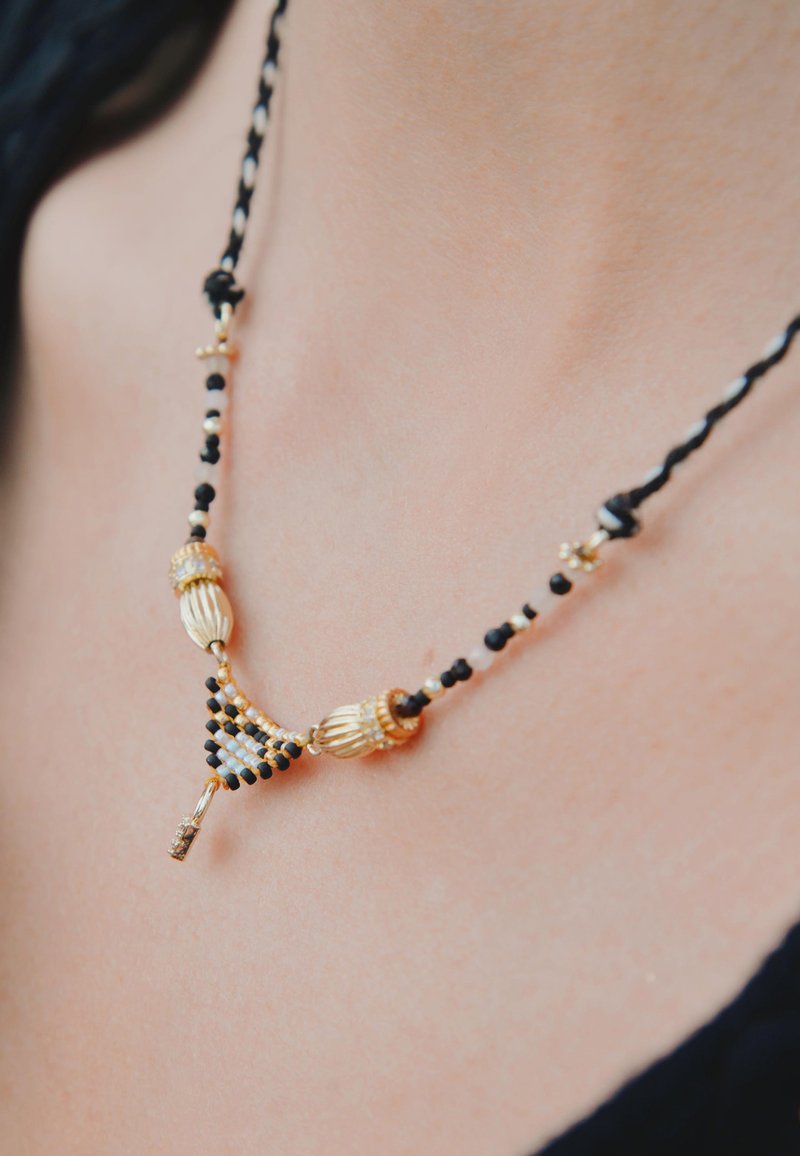 Collier de perles dorées et noires avec un design triangulaire, orné d'accents décoratifs et d'une corde tissée, mettant en valeur des motifs complexes.