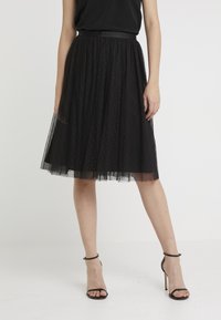 Gonna nera in tulle con un design a strati, caratterizzata da una cintura in raso e un motivo a pois. La modella indossa tacchi neri con cinturini.
