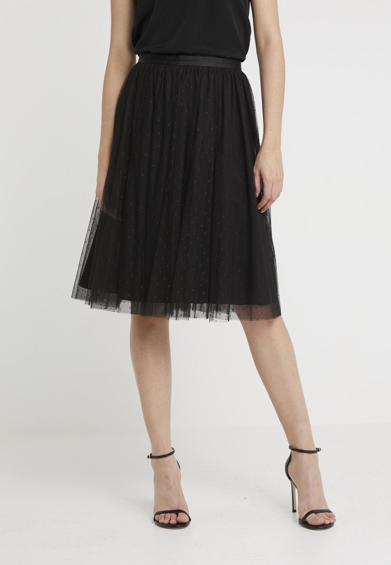 Gonna nera in tulle con un design a strati, caratterizzata da una cintura in raso e un motivo a pois. La modella indossa tacchi neri con cinturini.